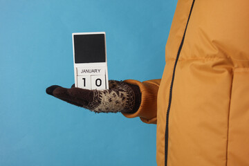 Man in yellow winter down jacket and gloves holding block calendar with date january 10 on blue...