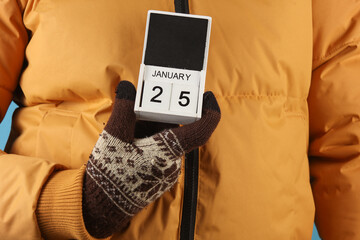 Man in yellow winter down jacket and gloves holding block calendar with date january 25 on blue...
