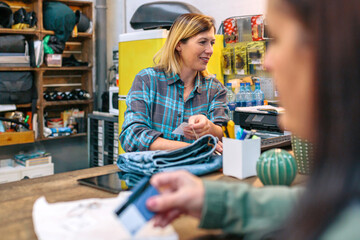 Cheerful young woman employee checking on computer the price of jeans while customer holding credit card to pay purchase in local store. Female shop assistant cashing clothing items to client.