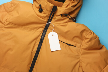 Yellow Winter down jacket with white blank tag on a string. Blue background. Shopping, sale concept. Template for design - obrazy, fototapety, plakaty