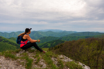 Naklejka na ściany i meble Young woman with backpack sitting on the mountain peak and beautiful mountains . Landscape with sporty girl, green grass, forest, hills, sky, sunbeams in fall. Travel. Nature