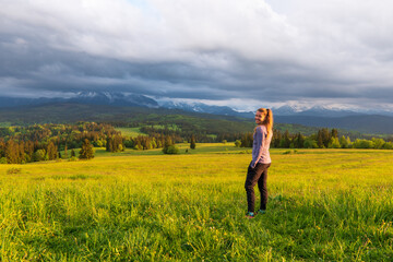 Young woman on the mountain peak with green grass and pink flowers looking at beautiful mountains in fog at sunset in summer. Colorful landscape with lying girl, forest, hills, sky. Travel and tourism