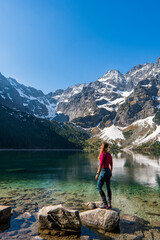 A tourist girl with a large backpack stands near an alpine lake in the stones in the lake Morskie oko or sea eye lake mountain against the backdrop of sea Eye and snow in autumn. - obrazy, fototapety, plakaty