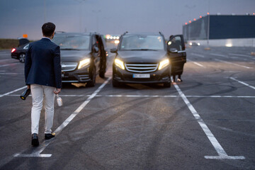 Businessman walks with alcohol drink to cars outdoors, having an informal celebration with partners on the parking lot
