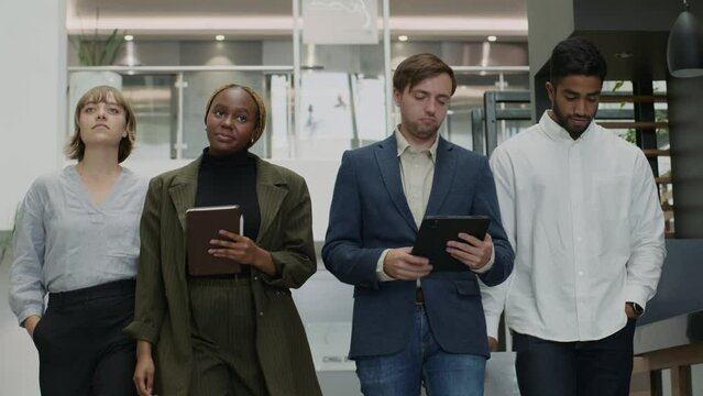 Four multiracial young adults in businesswear smiling while walking and talking in office