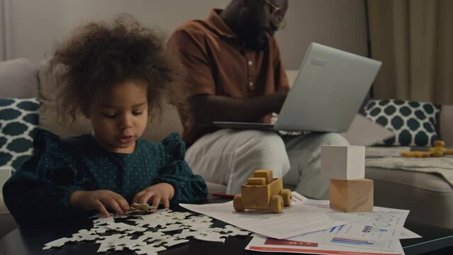 Black man working remotely, stressfully looking for his documents while his daughter playing with jigsaw puzzles