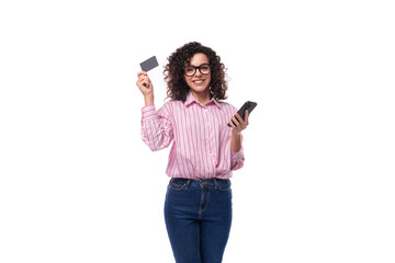 Fototapeta premium young european curly brunette lady in pink blouse holding plastic card mockup and smartphone on background with copy space