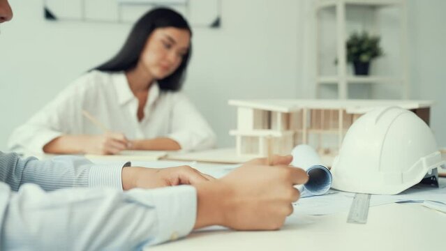 Professional architect engineer drawing blueprint and working together with young beautiful caucasian coworker on meeting table with safety helmet, house model, blueprint scatter around. Immaculate.