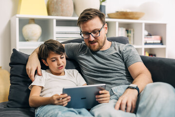 Front view of a Caucasian father and son, sitting on the couch and using digital tablet.