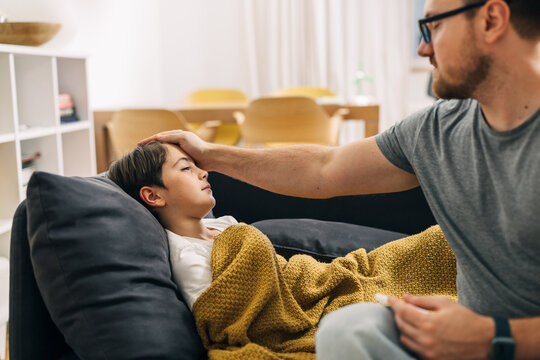 A young boy is laying sick on the sofa covered with a blanket, his fathers checks his temperature.