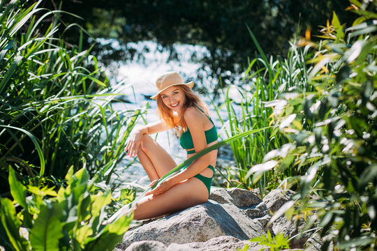 A slender laughing girl with blond hair and tanned skin, in a green bathing suit and with a hat, sits on stones in the grass, against the backdrop of a picturesque river in summer.