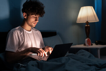 Young arabic man with laptop computer lying in bed at home at night, working or watch movies,...
