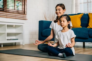 Fototapeten In a cozy home setting a young mother and her charming daughter practice family yoga in lotus position emphasizing mindfulness and meditation with smiles reflecting togetherness and happiness. © sorapop