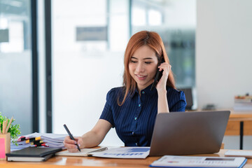 Obraz premium Young beautiful Asian woman using a smartphone and working with a laptop while sitting at an office desk, working from home concept.