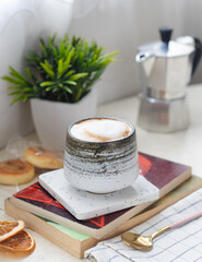 A cup of milk cappuccino coffee with pile of books on table with spoon and Italian moka pot, cookies in background 