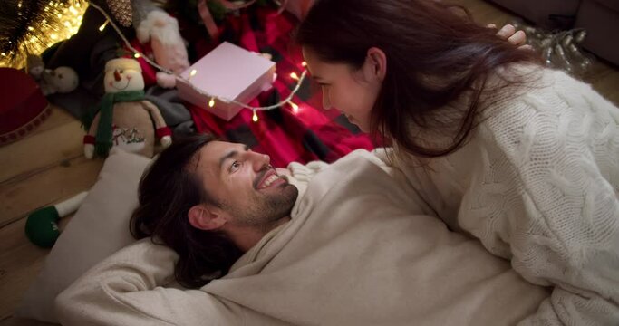 A brunette guy in a white sweater is lying on the floor on a pillow and a brunette girl is looking at him from above and lying next to him near New Year's toys and gifts in a cozy room in winter