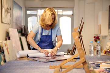 Confident focused female artist in blue apron standing at table and working with blueprints in her creative art studio