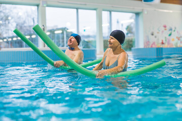 Smiling senior couple doing exercise in swimming pool with swim noodles. Happy retired people play...