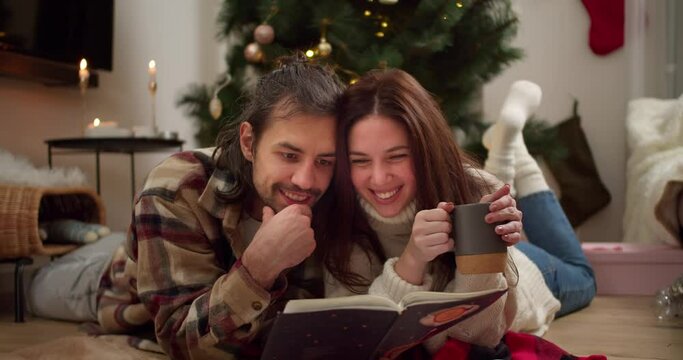 Interested guy and girl lying on the floor and reading a Christmas book along with a cup of hot drink in a cozy room with a Christmas tree and a decorated room in winter evening