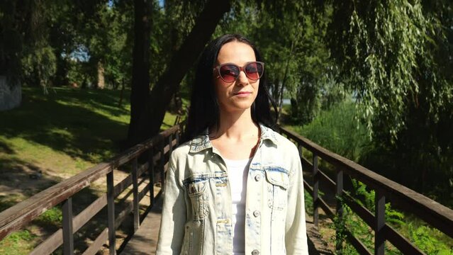 A young brunette woman in a denim jacket walks along a wooden bridge in a city park in summer. Slow motion portrait of a woman in sunglasses. A woman walks and looks around