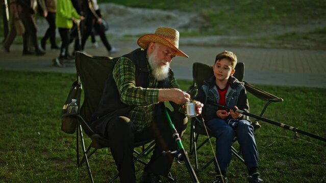 Grand dad showing his grandson how to put maggot on a fishing hook near riverside. Pensioner male teaching small grandchild catching fish together and sitting in chairs