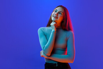 Answers and questions. Girl keeps chin on hand, fist and looking away pensively and think or dreaming against blue background in neon light.