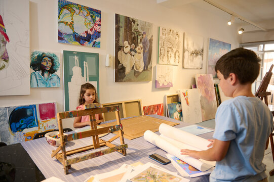 Adorable kids, a teenage boy and little child girl standing by a desk with pictures and drawing tools in the art class
