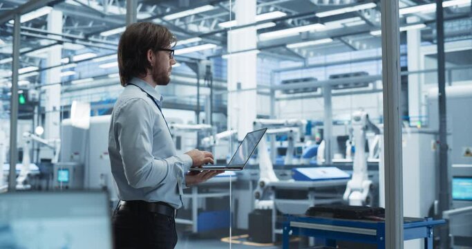 Factory Manager Working on Laptop at a Futuristic Industrial Manufacture with Autonomous Support Robots. Specialist Monitoring Technical Data on a Computer, Analyzing Reports and Planning Updates