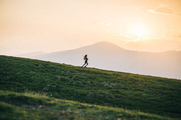 Woman trail running through alpine landscape in morning light, view of mountains behind her