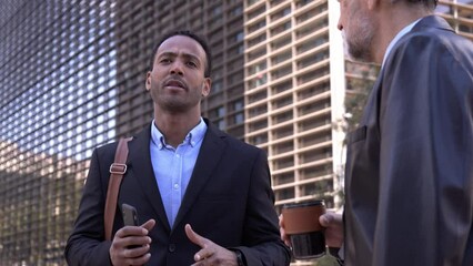 Two professional male of other nationalities with big age difference talk relaxed standing on city street on sunny morning. Diverse business men in formal suits together on break office work outdoors.