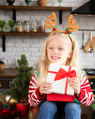 Happy child holding Christmas present in festive decorated kitchen. Kid in reindeer antlers with holly berries headband laughing and looking sideways