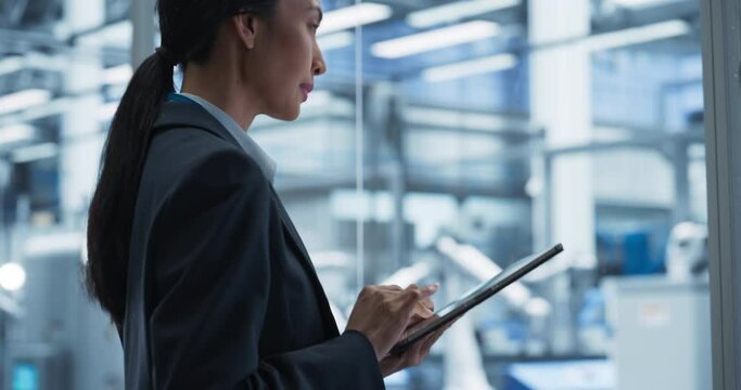 Beautiful Asian Female Engineer Standing Next to a Window in Office, Using Tablet Computer at an Electronics Manufacture. Specialist Using an Online Software to Program Robots on Assembly Line