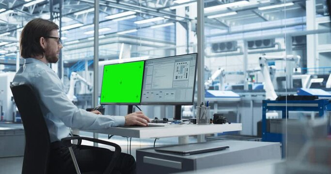 Young Software Developer Working on a Desktop Computer with Green Screen Chromakey Mock Up Display in a Factory Office. Technological Research and Development Plant with Automated Assembly Line