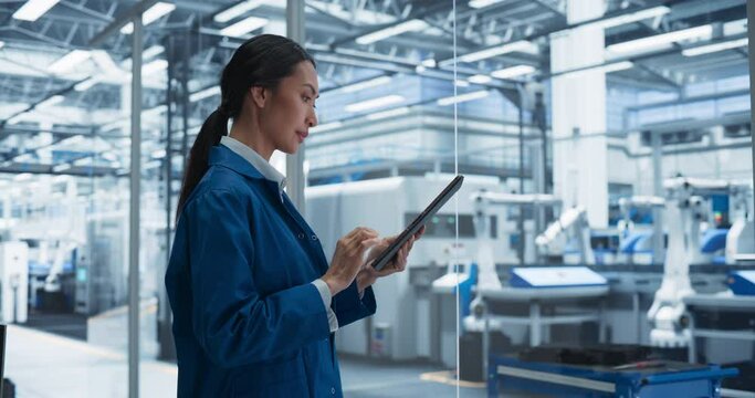 Focused Asian Engineer Monitoring and Analyzing Conditions at a Modern Electronics Factory on a Tablet Device. Facility with Automated Robot Hands Working with the Help of Artificial Intelligence
