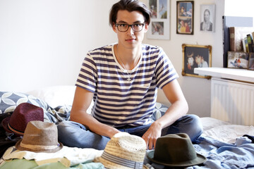 Fashion, hats and portrait of man in bedroom with fedora collection in home and apartment with nerd. Geek, style and person on bed with choice of vintage hat with trendy glasses and planning outfit - obrazy, fototapety, plakaty