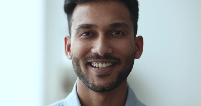 Serious handsome young Indian man getting happy, positive, cheerful, looking at camera with toothy smile. Face of attractive 20s guy with emotional changes. Close up facial portrait