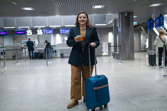 Smiling business woman going on business trip carrying suitcases while walking through airport passageway. Female holding passport with tickets and carrying suitcase while departing departure, copy