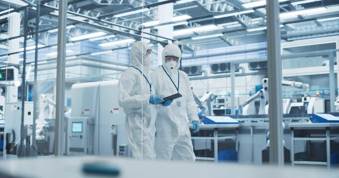 Factory Assembly Facility: Engineers in Coveralls and Masks Walking Through Workshop Facility and Using a Tablet Computer. In Background Robotic Arms Producing Modern High Tech Electronics Components