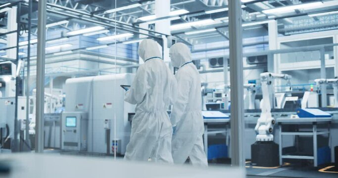 Sterile Modern Factory: Professionals in Coveralls Having a Conversation while Walking Through a Plant. Medical Electronics Manufacturing Laboratory with CNC Machinery and Automated Robotic Arms