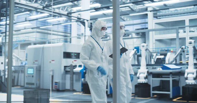 Scientists Walking in Full-Body Protective Coveralls, Masks and Goggles at a Manufacturing Plant. Specialists Using a Tablet Computer, Supervising Production at a Biotechnology Research Center