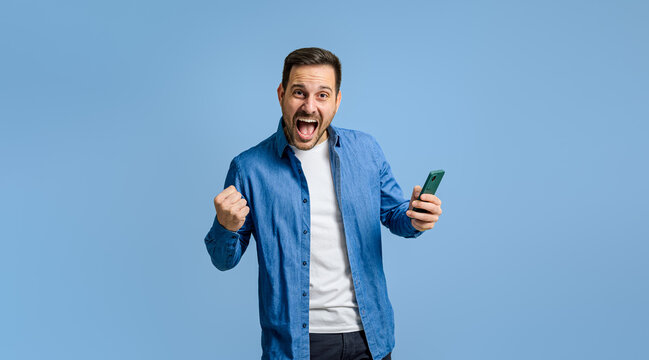 Businessman holding smart phone screaming and pumping fist ecstatically against blue background
