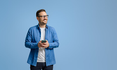 Smiling businessman holding mobile phone and looking away thoughtfully against blue background - obrazy, fototapety, plakaty