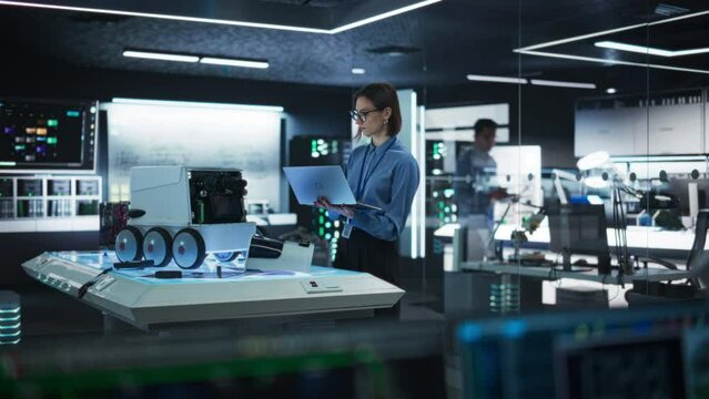 Portrait of a Female Technical Specialist Working in High Tech Research Laboratory with Modern Equipment: Female Developer Using Laptop to Write Code for an Advanced-Technology Delivery Robot