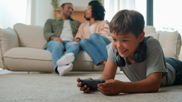 African American parents mother father talking on sofa little boy son child kid on floor play online video game on mobile phone internet addicted with smartphone happy multiracial family rest at home