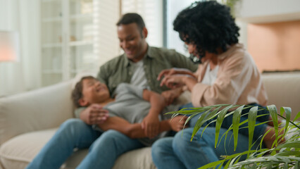 Bonding family African American parents with little adopted kid boy son tickling on home sofa...