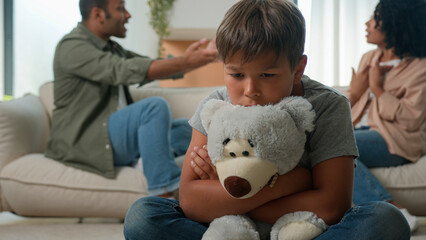 Upset Caucasian little boy preschool son hug teddy toy while African American woman mother and man...