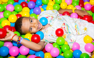 Fototapeta na wymiar Happy african black boy playing in ball pit on birthday party in kids amusement park and indoor play center. Child playing with colorful balls in playground ball pool.