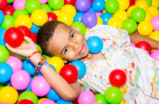 Happy african black boy playing in ball pit on birthday party in kids amusement park and indoor play center. Child playing with colorful balls in playground ball pool.
