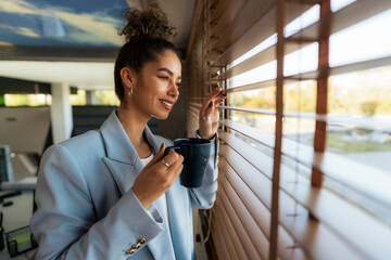 A beautiful young woman with curly hair, in her 20s, enjoys a coffee break in her modern office while gazing out the window.