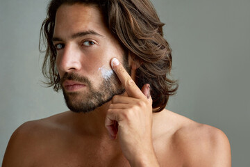 Portrait, facial or lotion with a shirtless man in studio on a gray background for his grooming routine. Skincare, face and beauty with the body of a young person with antiaging cream for his skin